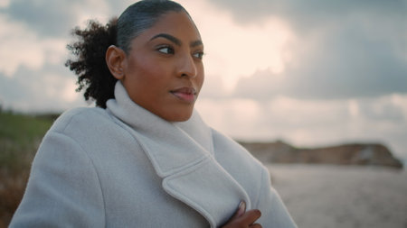 Closeup pensive african american rest cloudy shore. Black hair tourist get cold adjusting coat on autumn weekend. Thoughtful beautiful woman contemplating watching ocean. Lonely female take break.の写真素材