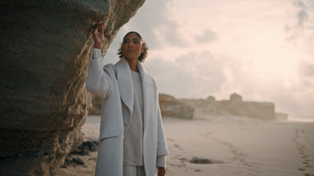 Beautiful girl touching cliff wall on empty beach. Serene african american enjoy quiet place admiring nature. Calm black hair model contemplating life look inspiration outdoors. Pensive woman restingの写真素材