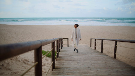 Lonely woman walking beach wooden pathway. Serene african american enjoy time at ocean shore. Black hair model strolling on cloudy day. Smiling beautiful girl taking break going peaceful place alone.の写真素材