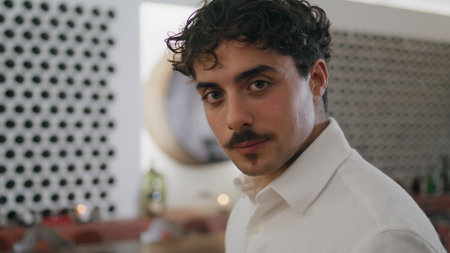 Portrait of attractive italian young man standing in winery cellar alone wearing white shirt. Curly mustached sommelier looking camera with light smile close up. Handsome guy relaxing in luxury bar.の写真素材