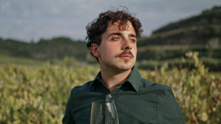 Young man winegrower looking at vineyard holding glass goblet with delicious white wine close up. Attractive italian winemaker watching grape plantation with harvest. Relaxed vintner enjoy vinery viewの写真素材