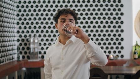 Portrait of italian sommelier drinking red wine from elegant glass standing at winery restaurant with bottle collection on backdrop. Attractive winemaker looking camera enjoying grape alcohol close upの写真素材