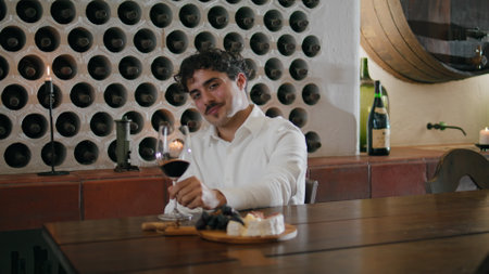 Young attractive professional wine taster looking on glass with swirling red alcohol drink close up. Confident curly sommelier shaking wineglass mixing grapevine inside evaluating color at degustationの写真素材