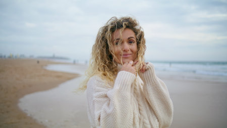 Woman posing autumn beach in warm sweater. Carefree blonde enjoying ocean shore relaxing on romantic date. Beautiful stylish girl looking camera feeling satisfied with life. Calmness happiness conceptの写真素材