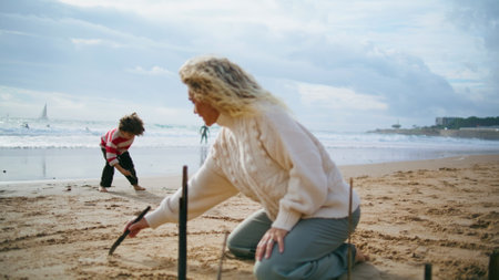 Family writing beach sand together. Creative mother son resting at ocean shore playing games on autumn holidays. Focused foster mom adopted child learning enjoying weekend. Playful childhood concept.の写真素材