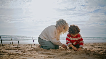 Mother relaxing autumn beach with little son. Carefree family building sand castle enjoying weekend at ocean shore. Curly single mom nanny spending quality time with child. Family games creativity.の写真素材