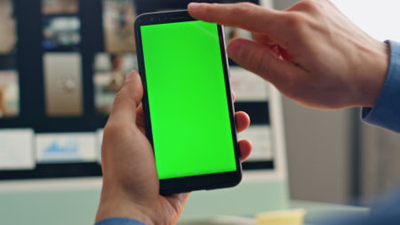 Businessman hand swiping mockup smartphone at table closeup. Unknown manager holding chromakey telephone watching social media at office. Man finger tapping display relaxing with greenscreen cellphoneの写真素材