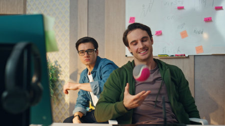 Creative colleagues working computer in office closeup. Relaxed smiling businessman playing ball listening partner at hipster workplace. Two designer guys looking monitor communicating together indoorの写真素材
