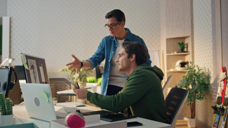 Smiling colleagues working computer in corporate office. Eyeglasses man giving advices to partner. Two guys looking pc monitor screen enjoying friendly conversation. Active creators having deadlineの写真素材