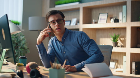 Positive creator talking cellphone at cabinet. Close up smiling designer watching computer screen gesturing hand at office. Creative business man consulting colleague holding mobile phone indoorsの写真素材