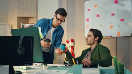 Serious man working computer looking screen at business cabinet. Friendly colleague giving coffee to office partner. Smiling coworkers holding hot beverages staring monitor together. Team work conceptの写真素材