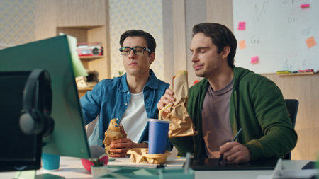 Involved partners tasting food discussing job at business place. Closeup two guys having lunch break talking. Friendly designers creating ideas enjoying aromatic sandwiches at office. Work lifestyleの写真素材