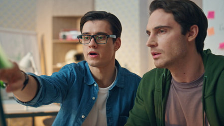 Focused colleagues pointing computer in office closeup. Eyeglasses man giving advices to partner. Two guys looking pc monitor screen working on project together. Serious creators having deadlineの写真素材
