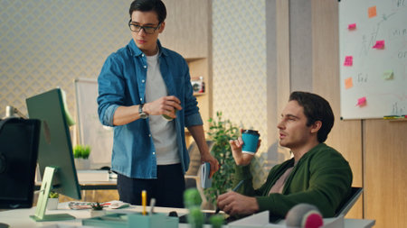 Two creators discussing project at sunny office. Eyeglasses smart man saying opinion going to break. Focused freelancer drinking coffee looking computer screen attentively. Designer involved in workの写真素材
