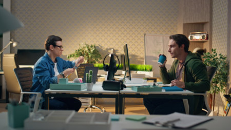 Creative designer showing swatches to colleague. Inspired creators communicating together at modern office. Focused man listening partner drinking coffee at computer workplace. Business team speakingの写真素材