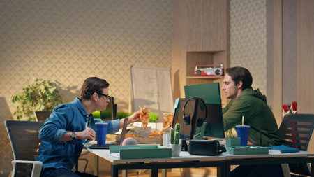 Hungry startupers taking pizza slices at work place. Two young men enjoying tasty snack food at break together. Friendly male colleagues sharing corporate lunch using food delivery service at officeの写真素材