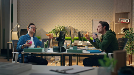Talking creators eating pizza together for lunch. Hungry designers communicating workplace interior. Relaxed men enjoying dinner meal speaking at office table. Smiling colleagues having takeout foodの写真素材