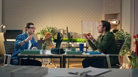 Office workers eating lunch at monitor table informal office. Brunette freelance guys enjoying relaxed communication talking. Smiling cheerful teammates having takeaway food meal at modern workplaceの写真素材