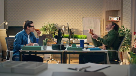 Excited partners tasting food joking at open space. Two laughing guys enjoying lunch break talking together. Friendly designers having fun eating aromatic pizza at office. Corporate work lifestyleの写真素材
