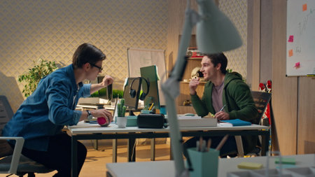 Young businessmen working office communicating together. Eyeglasses man asking question to smiling colleague. Web designer peeping computer screen at creative workplace. Coworkers exchanging ideasの写真素材