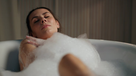 Relaxed woman touching skin in foam bath closeup. Groomed naked girl taking bathtub closed eyes at home. Romantic brunette lady feeling lazy enjoying evening beauty treatment alone. Hygiene conceptの写真素材