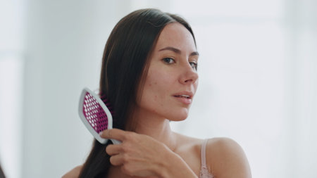 Relaxed girl domestic hairstyling close up. Peaceful lady combing silky hair looking mirror reflection at bathroom. Smiling attractive woman brushing chevelure in morning. Everyday beauty routineの写真素材
