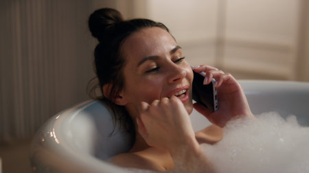 Romantic model talking smartphone chilling bathtub closeup. Relaxed happy girl laying hot bubbles filled bath flirting cellphone. Smiling woman enjoying spa procedures at home. Domestic beauty routineの写真素材