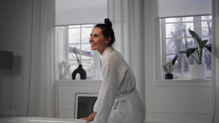 Happy girl sitting bathtub smiling at evening home. Satisfied woman enjoying beauty routine. White gown cheerful lady relaxing alone. Positive model preparing to foamy bath having spa proceduresの写真素材