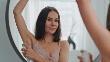 Smiling woman applying antiperspirant at home closeup. Cute lady spraying armpits deodorant enjoying daily procedures. Happy girl looking mirror reflection. Body care daily hygiene routine conceptの写真素材