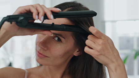 Brunette lady straightening hair in bath room portrait. Smiling model using iron equipment at home closeup. Groomed woman looking camera styling long chevelure pov. Morning haircare routine conceptの写真素材