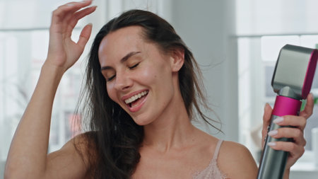 Portrait excited lady hair drying at light windows room. Close up emotional woman making hair style in white bathroom. Long haired model holding hairdryer at home. Morning beauty treatments conceptの写真素材