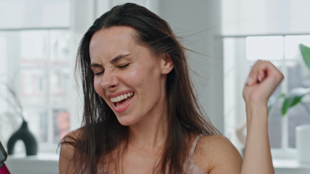 Excited girl drying hair at bathroom portrait. Overjoyed woman looking camera dancing with dryer. Close up happy model touching windy hairstyle singing at windows apartment. Female beauty routineの写真素材