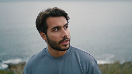 Calm bearded man walking on gloomy sea shore alone close up. Handsome young guy strolling on cloudy nature coast. Relaxed attractive tourist enjoy overcast weekend on beach. Vacation trip concept.の写真素材