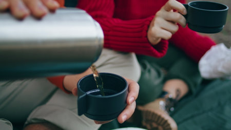 Man hands pouring drink from metal  to cup close up. Unknown guy filling mug by hot tasty beverage on picnic outdoors. Unrecognizable couple tourists sitting blanket enjoy weekend on nature.の写真素材