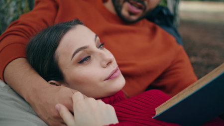 Tender woman relaxing at picnic lying on man chest closeup. Bearded boyfriend reading book aloud leaning on park tree. Romantic couple enjoy weekend on nature together. Tranquil family leisure conceptの写真素材