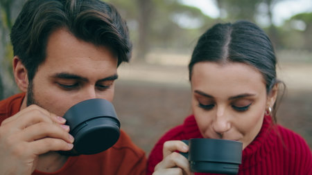 Loving cute couple drinking hot beverage at picnic on autumn nature close up. Portrait of relaxed smiling man woman holding cups enjoying coffee in park. Romantic pair tourists relax together outdoorsの写真素材