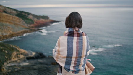 Back view relaxed young woman standing in front gray calm ocean. Unknown carefree girl looking on beautiful evening seascape. Dark haired peaceful lady relaxing at scenic sea shore wrapped in plaid.の写真素材