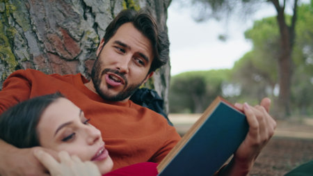 Handsome bearded man reading book at picnic for relaxed woman close up. Attractive calm girl lying on boyfriend chest enjoy weekend at nature. Cute romantic couple spending vacation together in park.の写真素材