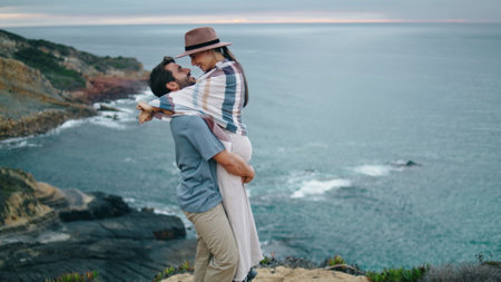Loving young couple hugging on beautiful cloudy coast hill. Attractive bearded man lifting spinning smiling woman near calm ocean. Happy girl in hat kissing boyfriend at nature. Honeymoon love conceptの写真素材