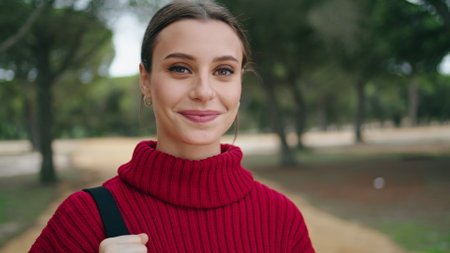 Portrait of cute young woman standing in front beautiful green forest wearing red sweater. Attractive happy girl holding bag going to walk on nature close up. Pretty brunette smiling looking camera.の写真素材