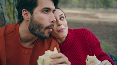 Couple relaxed tourists eating in forest enjoying beautiful nature view close up. Calm carefree family tasting sandwiches sitting outdoors. Attractive woman putting head on man shoulder feeling happy.の写真素材