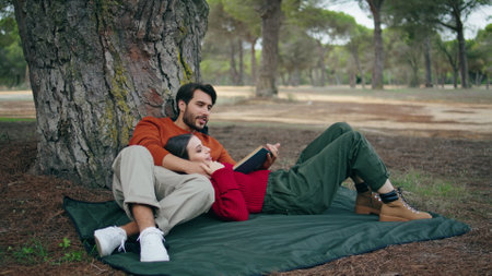 Tender couple lying on blanket at romantic forest picnic. Attractive bearded man reading loud book leaning on tree. Carefree young woman relaxing on boyfriend chest outdoors. Family leisure concept.の写真素材