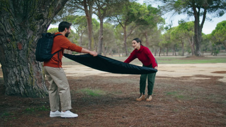 Happy young couple spreading blanket on yellow grass autumn forest. Cute positive pair preparing place for picnic on nature. Two active travelers with backpack going to rest on soft plaid together.の写真素材