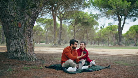Young carefree couple having picnic in autumn park sitting at blanket. Attractive bearded man giving cup beverage to beautiful woman. Happy smiling family enjoy romantic weekend on nature together.の写真素材