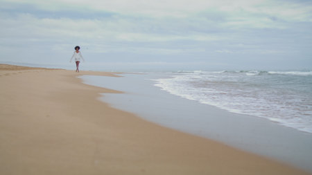 Slim girl walking ocean shore on gloomy day. Relaxed african american woman rest enjoying calm sea. Romantic tourist go sandy coastline in white bikini. Worried person contemplating personal issues.の写真素材