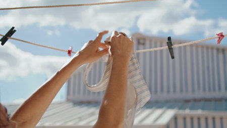 Closeup hands hanging bra at beach house. Stylish striped beachwear air drying on sunny day. Unrecognized woman tourist clipping wet swimsuit laundry after pool. Summertime vacation rest at seashoreの写真素材