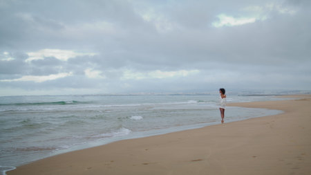 Dreamy woman walking ocean beach on cloudy day. Romantic african american girl enjoying sea coastline going barefoot in white outfit. Attractive black haired lady relaxing contemplating at shore wavesの写真素材