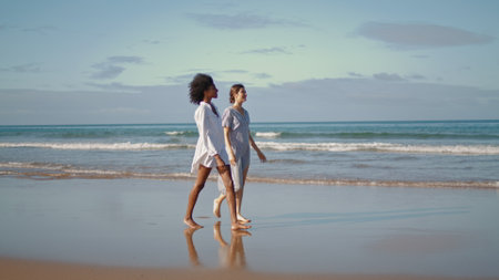 Lgbt couple go ocean coast in sunlight. Carefree girlfriends enjoy conversation walking sandy beach on summer weekend. Smiling multiethnic women sharing news at sea shore. Girls friendship concept.の写真素材
