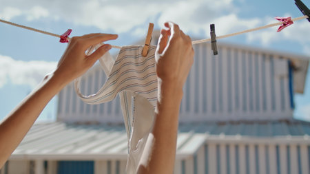Woman hands taking swimsuit off rope closeup. Striped bikini bra air drying in summer sunlight. Unrecognized girl tourist removing underwear from dryer in vacation house. Beachwear lingerie concept.の写真素材