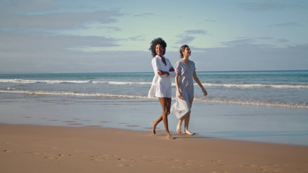 Happy women enjoying ocean walk. Smiling lesbian couple resting sandy beach talking on summer vacation. Beautiful multiethnic girls partners spending weekend at shore. Carefree wonderful life concept.の写真素材